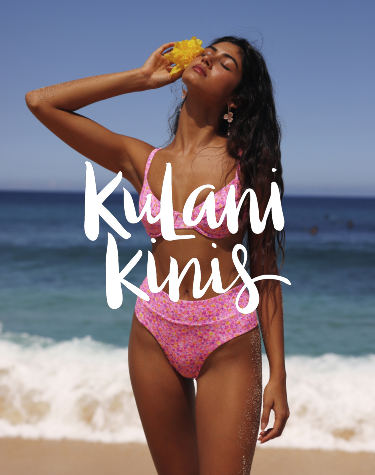 Woman strolling along the beach in a vibrant pink floral bikini from the Kulani Kinis brand, with a delicate flower tucked in her hair, embracing a tropical vibe.