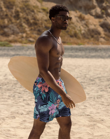 Man standing on the beach holding a small surfboard, wearing a swimsuit with a tropical foliage print in vibrant blue and pink tones, ready for a seaside adventure.