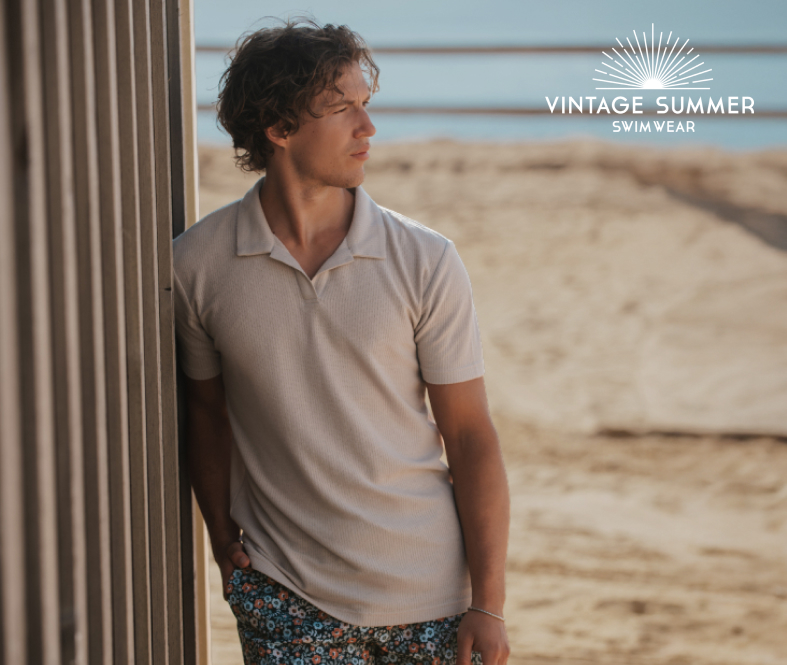 Man standing on the beach wearing a beige T-shirt paired with short swim trunks, enjoying a casual seaside moment.
